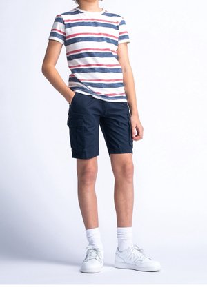 Dziecko stoi w paskowanej niebiesko-białej i czerwonej koszulce z krótkim rękawem, niebieskich szortach cargo, białych skarpetkach i białych trampkach na jednolitym tle.