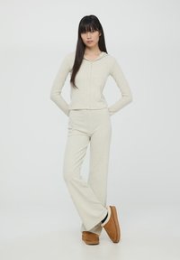 Jasno beżowy, prążkowany zestaw wypoczynkowy z kapturem na zamek i spodniami dzwonami. Uzupełniony o brązowe kapcie z puszystym wyściółką.