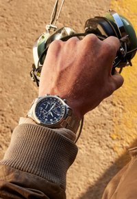 Stalowy zegarek chronograf z niebieską tarczą, białymi cyframi i srebrnymi akcentami, noszony na mankiecie swetra o teksturowym splocie.