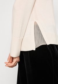 Pull beige à manches longues avec fente latérale porté sur un sous-vêtement côtelé, associé à un pantalon en velours noir, montrant une main détendue le long du corps.