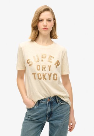 Crème t-shirt met "SUPER DRY TOKYO" in metallic gouden letters. Korte mouwen, losse fit, gecombineerd met blauwe jeans. Minimalistisch ontwerp.
