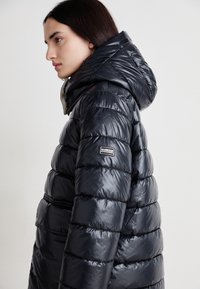  casaco puffer preto com gola alta e capuz, design acolchoado e textura brilhante. Branding notável no ombro esquerdo.