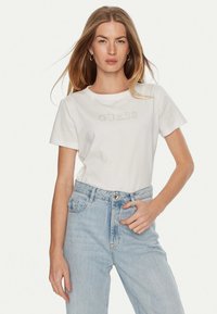 T-shirt bianca in cotone con maniche corte e colletto rotondo, con logo "GUESS" in rilievo. Abbinata a jeans in denim azzurro chiaro a vita alta.