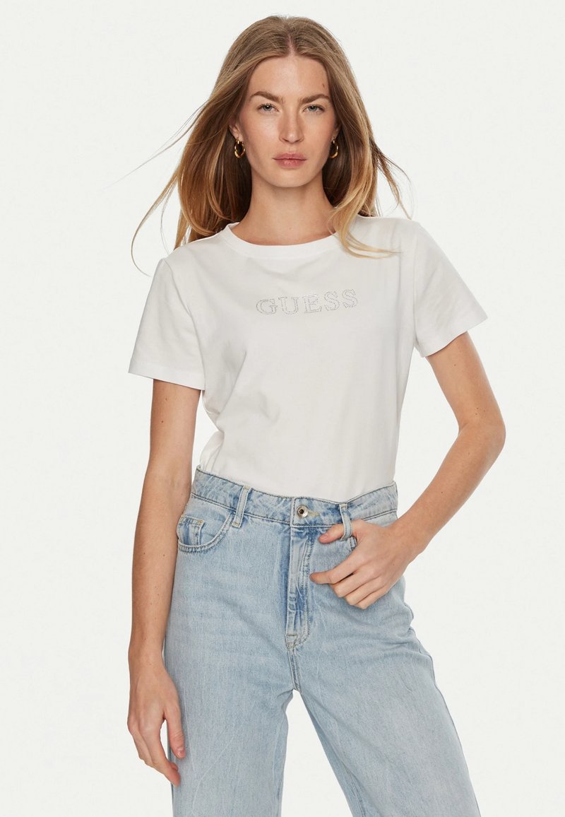 T-shirt bianca in cotone con maniche corte e colletto rotondo, con logo "GUESS" in rilievo. Abbinata a jeans in denim azzurro chiaro a vita alta.