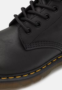 Stivaletto in pelle nera con punta tonda, lacci neri e cuciture gialle lungo la suola. Presenta passanti e superficie testurizzata.