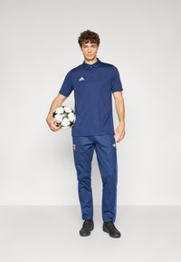 Polo azul-marinho com gola e logótipo, combinado com calças de treino azul-marinho com riscas laterais azuis e sapatos de futebol pretos, segurando uma bola de futebol.