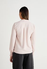 Helles rosa Langarmbluse mit rundem Kragen, gerippten Manschetten und einem Rückenfalz, kombiniert mit schwarzen, gestreiften Hosen. Glatte Textur.