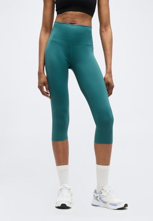 Nő, aki türkizkék magas derekú, rövidített leggingset, fekete sportmelltartót, fehér zoknit és fehér sportcipőt visel, egyszínű háttér előtt áll.