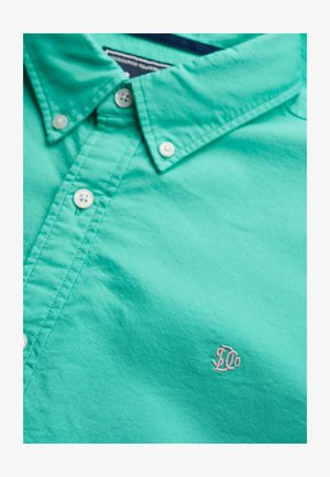 Chemise turquoise à boutons, en coton. Dotée d'un col standard, de boutons blancs et d'un petit logo rose sur la poitrine. Texture lisse.