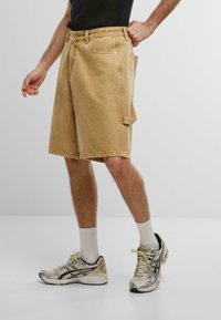 Pantalones cortos de mezclilla beige con un bolsillo lateral, un corte recto y detalles de costura visibles, combinados con zapatillas deportivas plateadas y negras.
