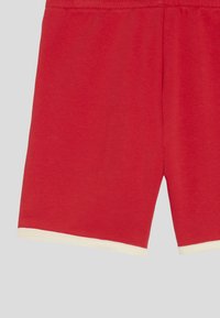 Rode shorts met witte bies aan de zoom en zichtbare elastische tailleband, weergegeven op een effen lichtgrijze achtergrond.