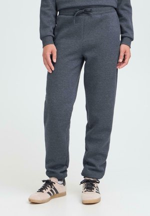 Mann med mørkegrå joggebukser med snøring og beige sneakers med svarte striper, stående på en lys bakgrunn.