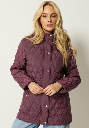Femme blonde portant une veste matelassée violet foncé avec des boutons-pression argentés, debout devant un fond clair uni.