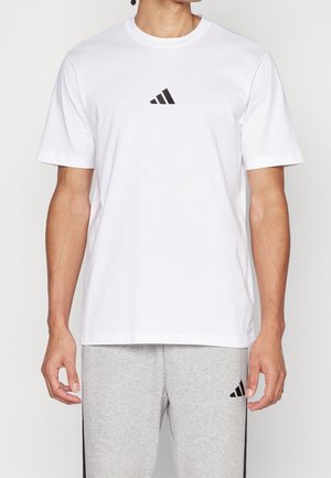 Mężczyzna ubrany w białą koszulkę Adidas i szare spodnie dresowe z czarnymi logo Adidas, stojący na jednolitym tle.