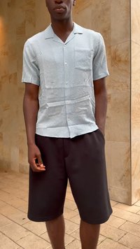 Homem com uma camisa de botão de mangas curtas azul claro texturizada e calções pretos até aos joelhos, de pé contra uma parede de azulejos bege.
