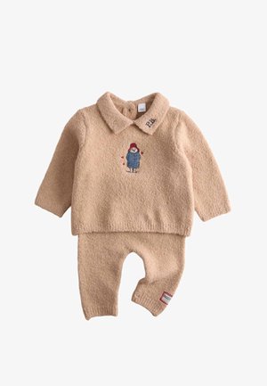 Blødt beige babyoutfit med lange ærmer, krave, broderet bjørn i blå jakke på brystet og rød lap på venstre benmanchet.
