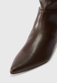 Smeđe kožne gležnjače s aristokratskim vrhom, glatkom površinom i minimalnim detaljima šivenja. Potplat je ravan s blagom fleksibilnošću.