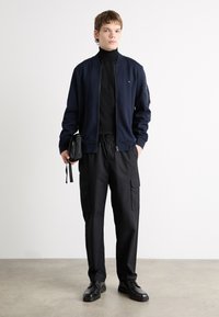 Jūras spēku bumbvedēju jaka ar rāvējslēdzēju, melns turtlenecks, melni kravas bikses ar kabatām, melnas kurpes un maza melna soma. Gluda tekstūra.