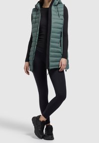 Osoba w turkusowej, pikowanej kamizelce bez rękawów, na czarnej bluzce z długim rękawem, czarnych legginsach i czarnych butach sportowych, stojąca z nogami skrzyżowanymi.