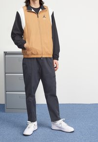 Ruskeankeltainen ja musta vetoketjullinen takki, jossa on valkoinen paneelisuunnittelu; yhdistetty tummien housujen ja valkoisten lenkkareiden kanssa. Takissa on logo.