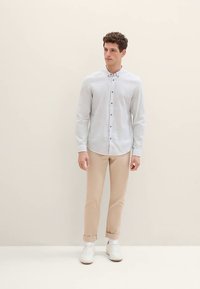 Ljusblå skjorta med lång ärm och knappar i mörk färg, kombinerad med beige avsmalnande byxor och vita sneakers. Slät tygstruktur, enkel design.