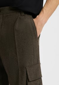 Bruna herringbone cargobyxor tillverkade av textilier med strukturerad yta och sidofickor. Har en avslappnad passform och synliga plisseringar fram.