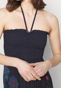 Havsblå klänning utan axelband med smockad midja och volangkant, justerbara remmar och blommönster broderat på kjolen.