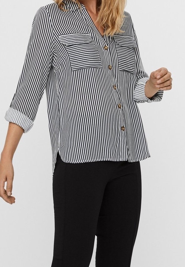 Femme portant une chemise à rayures verticales noires et blanches à boutons, manches retroussées, et un pantalon noir ajusté, sur un fond clair uni.