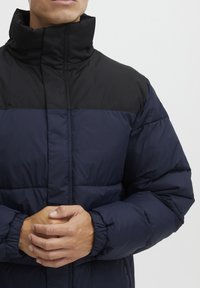 Paduotas švarkas su juodu viršutiniu skyriumi ir tamsiai mėlynu apatiniu skyriumi, aukštu vvišdelio, elastingomis rankovėmis ir siuvinėtu raštu.