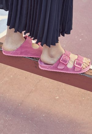 Sandales en suède rose avec deux boucles ajustables, design ouvert, et semelle texturée, posées sur un rebord à côté d'une jupe noire fluide.