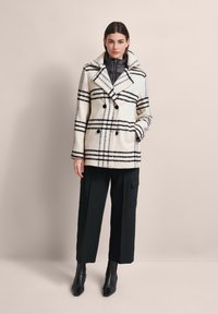 Doppelreihiger Wollmantel in Creme mit schwarzem Karomuster, großem Kragen und zwei Vorderstaschen; kombiniert mit schwarzen weit geschnittenen Hosen und Stiefeletten.