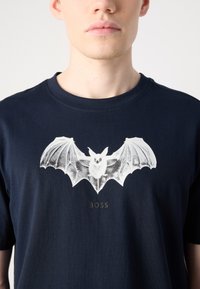 Námořnická modrá bavlněná tričko s bílým potiskem netopýra a slovem "BOSS" v šedé barvě pod designem. Kulatý výstřih, krátké rukávy.