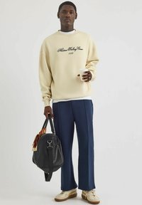 Sudadera crema con texto negro, pantalones azul marino y zapatillas beige; sosteniendo una taza de café, con una bolsa negra con un detalle estampado.
