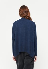 Sötétkék, oversized pulóver magas nyakú, ejtett vállú és hosszabb hátrésszel. Puha, textúrált anyagból készült.