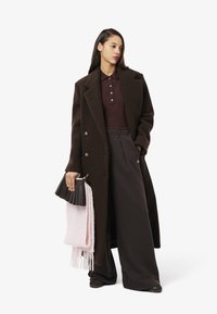 Manteau en laine marron, polo bordeaux avec boutons, pantalon noir large, tenant un sac à main noir texturé et une écharpe rose à franges.