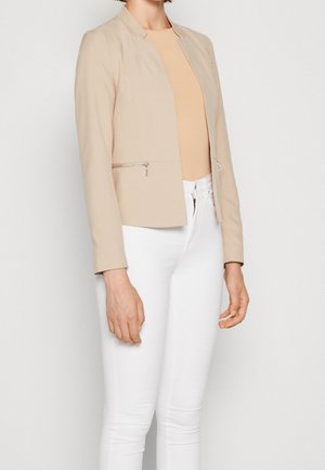 Femme portant un blazer beige par-dessus un haut marron clair et un pantalon blanc, debout devant un fond blanc uni.