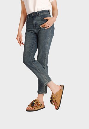 Donna con jeans blu e sandali slip-on beige con perline colorate e frange, in piedi davanti a uno sfondo chiaro e uniforme.
