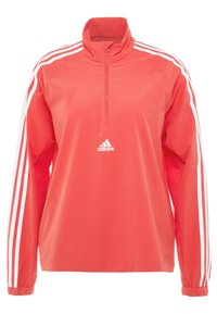 Punainen villapusero takki, jossa on valkoiset raidat hihoissa, korkea kaulus ja puoliväli vetoketju edessä. Keskellä on Adidas-logo. Silkkinen tekstuuri.