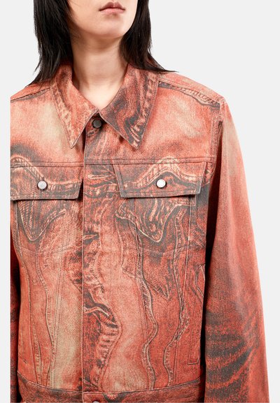 Chaqueta de mezclilla roja con un diseño abstracto, dos bolsillos en el pecho y botones de presión plateados. La textura parece ligeramente desgastada o descolorida.