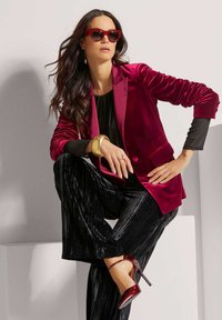 Dieprode fluwelen blazer met geplooide mouwen, gecombineerd met een zwarte top en geplooide zwarte broek. Rode puntige hakken en bijpassende zonnebril.