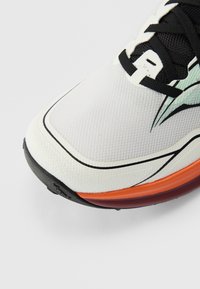 Λευκό αθλητικό παπούτσι με υφασμάτινο επάνω μέρος, μαύρα κορδόνια και πολύχρωμη σόλα με πορτοκαλί και μωβ πινελιές.