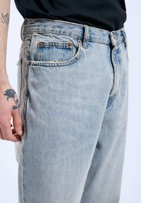 Jeans de mezclilla azul claro con un dobladillo deshilachado, remaches de cobre y un bolsillo trasero. Se muestran en un lado de un modelo masculino con un tatuaje visible en la mano.