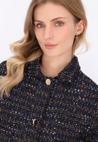 Una giovane donna con capelli biondi ondulati sorride, indossando una giacca blu navy con texture e macchie colorate e bottoni dorati, su uno sfondo bianco.