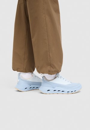 Osoba nosící světle modré tenisky s tlustými podrážkami a hnědé ohrnuté kalhoty na jednoduchém pozadí.