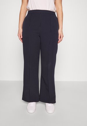 Personne portant un pantalon large rayé bleu marine et des baskets blanches, debout devant un fond clair uni.