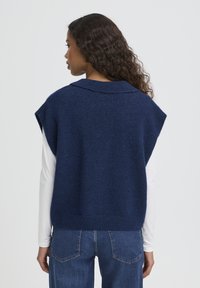 Pull sans manches en tricot bleu marine avec un large col. Texture douce, bordure côtelée, et coupe décontractée. Porté sur une chemise blanche à manches longues.
