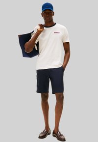 T-shirt bianca a maniche corte con colletto blu navy e logo, pantaloncini blu navy, cappellino blu, che tiene una borsa a tracolla blu navy con dettagli crema. Mocassini marroni.