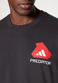 Črna bombažna majica s kratkimi rokavi, ki ima rdeč logotip s tremi belimi črtami in besedo "PREDATOR" spodaj, ter okroglo ovratnico.
