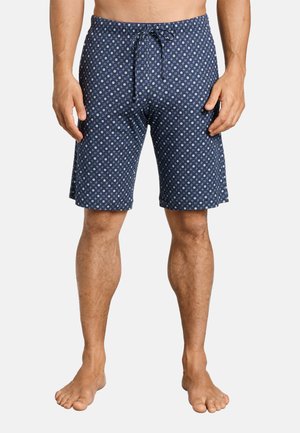 Modello maschile che indossa pantaloncini blu navy fino al ginocchio con un motivo geometrico bianco, in piedi a piedi nudi su uno sfondo semplice.