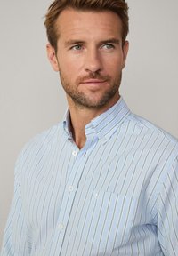 lichtblauw shirt met knopen, voorzien van verticale witte strepen, button-down kraag, één borstzak en een subtiel logo op de zak.